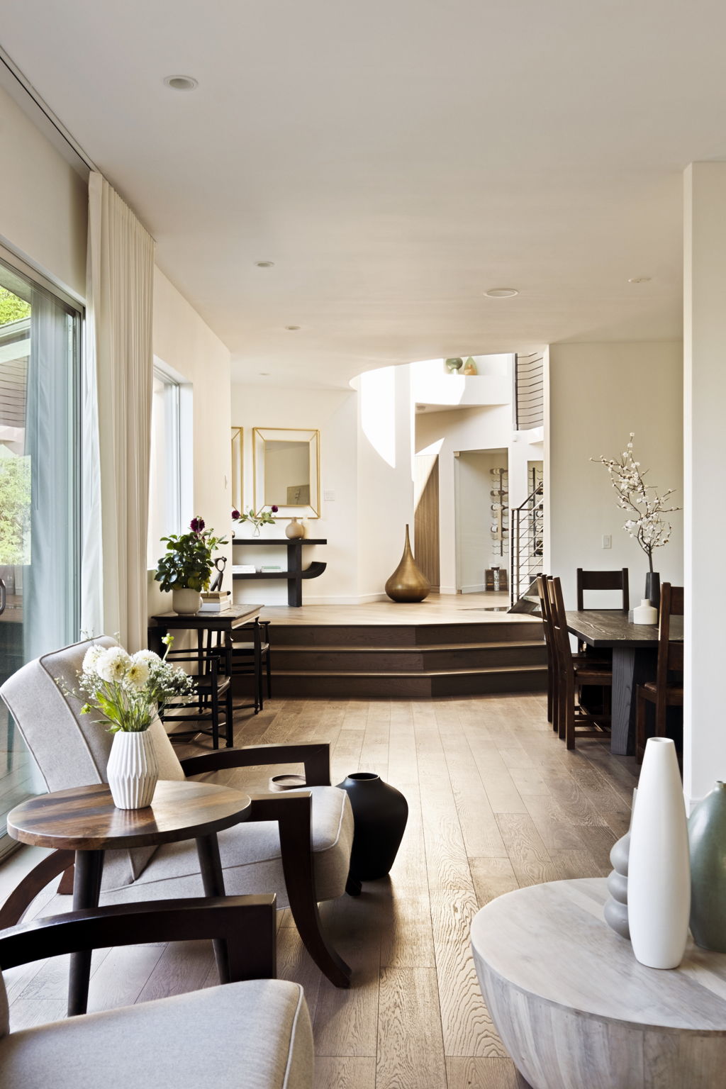 Modern living and dining area with large windows, light wood floors, and broad steps to a raised sitting area.