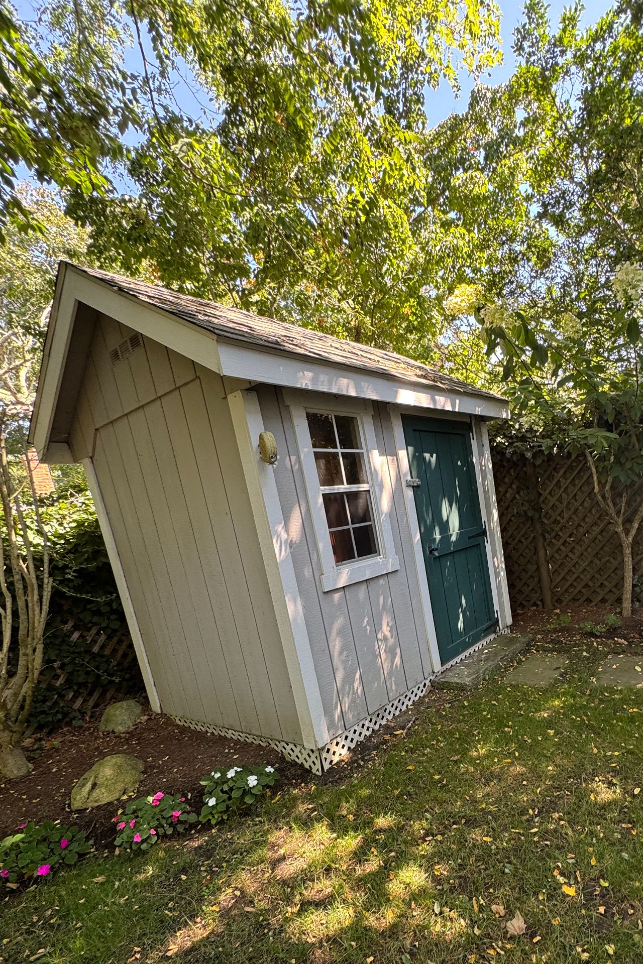 Small gray garden shed with a green door beneath leafy trees.