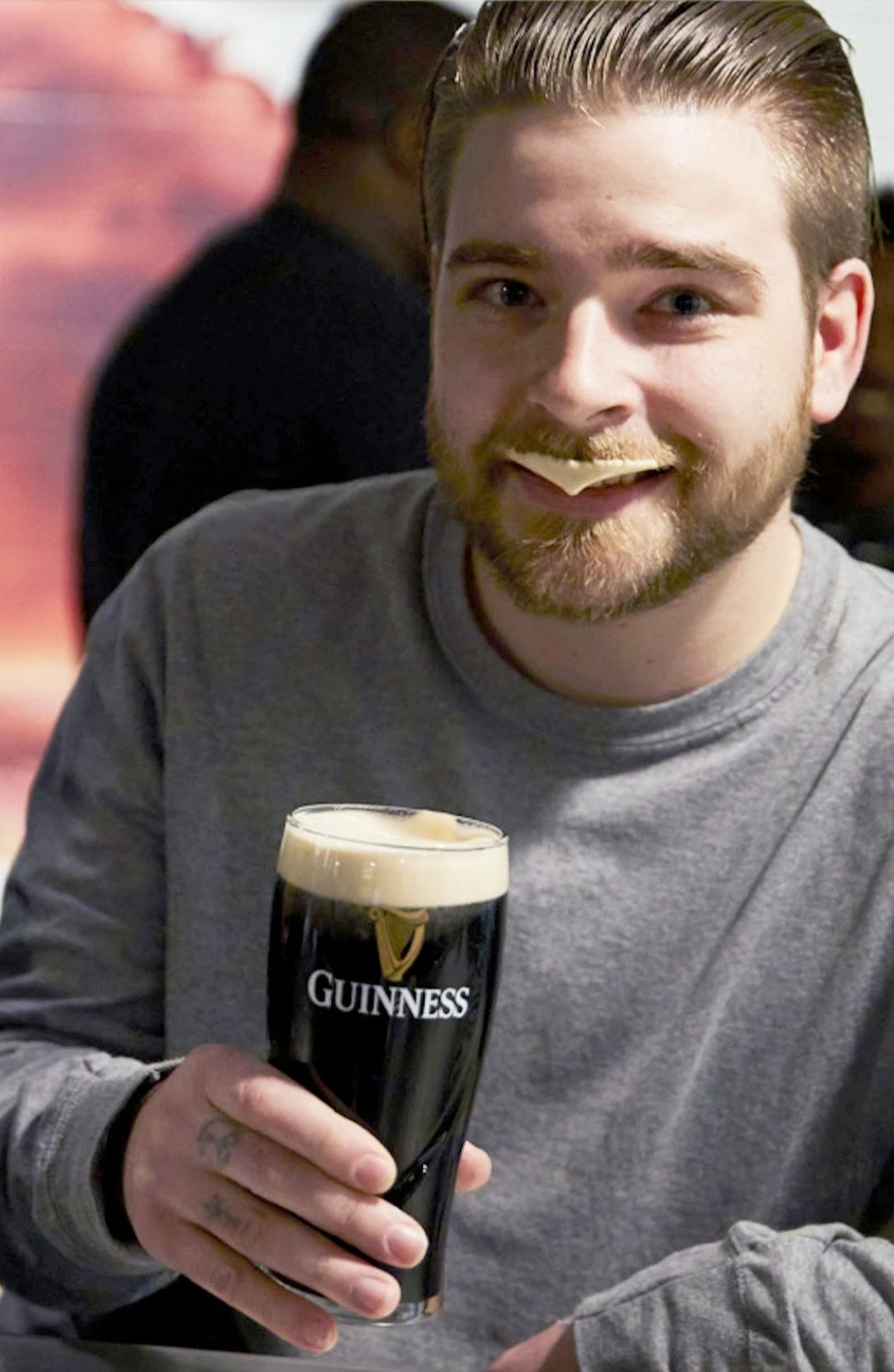 Bearded man holding a pint of Guinness with a small snack resting on his mustache.