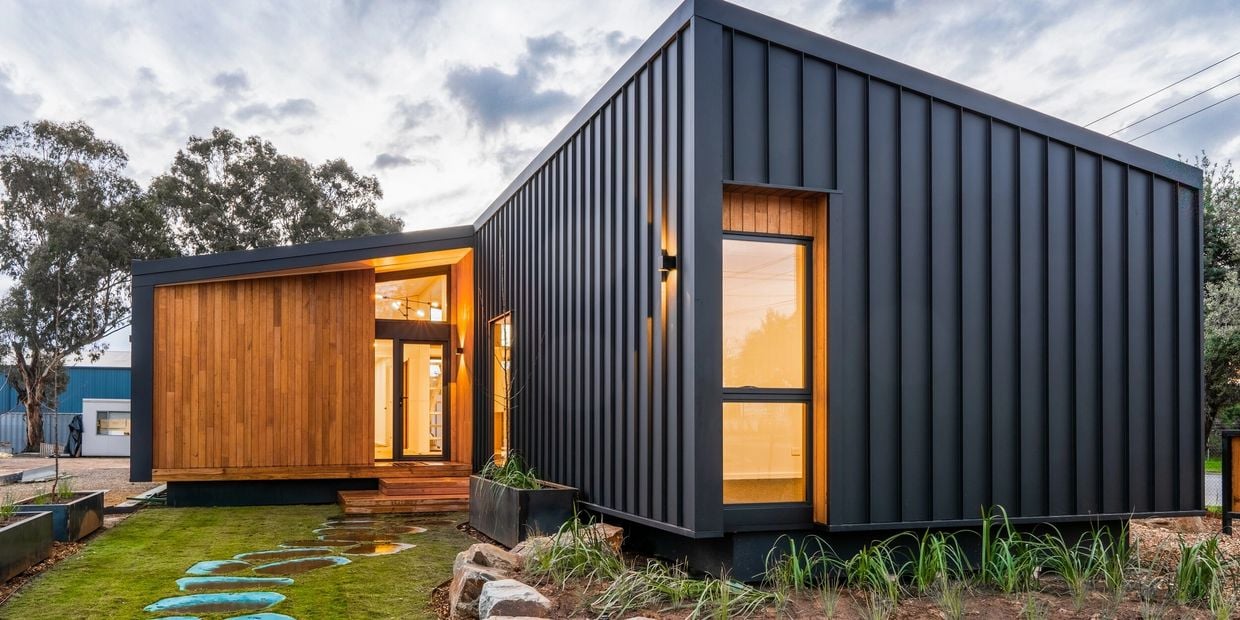 Modern modular home with dark metal siding, a wood-clad entrance, and stepping stones across the lawn.