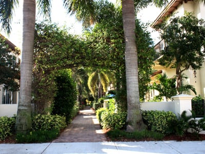 Brick walkway through landscaped residential grounds, framed by palm trees and greenery.