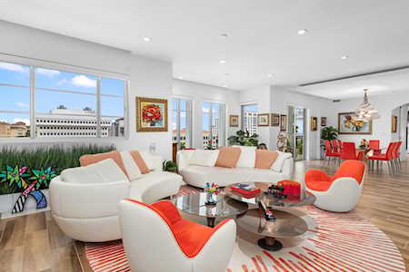 Contemporary living room with orange-accented white seating, large windows, and an adjoining dining area.