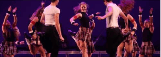 Dancers perform onstage in plaid skirts under purple lighting.