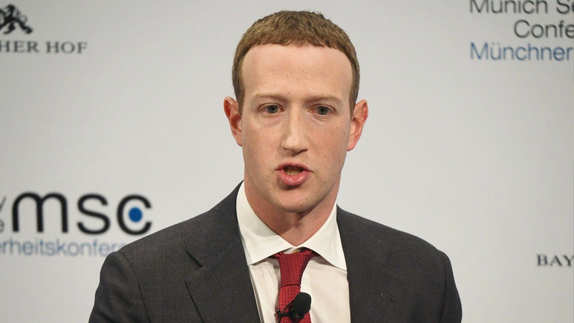 Mark Zuckerberg speaking at a conference in a dark suit and red tie.