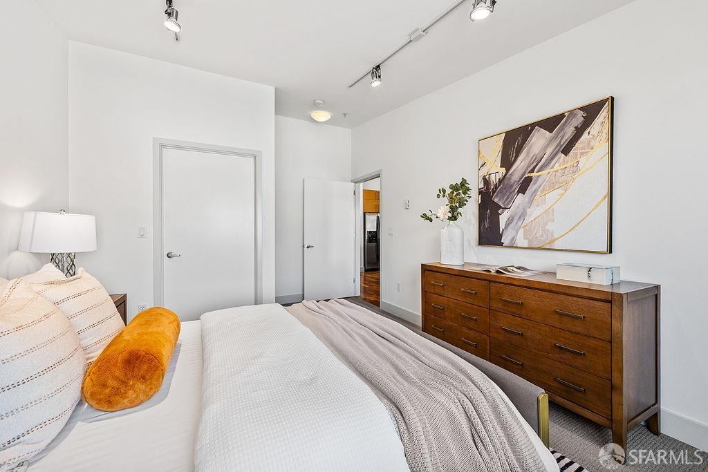 Furnished bedroom with a white bed, orange accent pillow, and wooden dresser beneath abstract artwork.