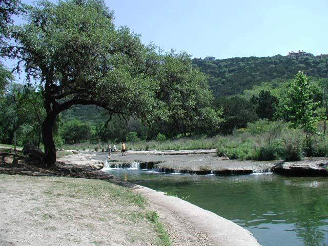 A creek and low waterfall beside a shaded trail, with two people near the water.