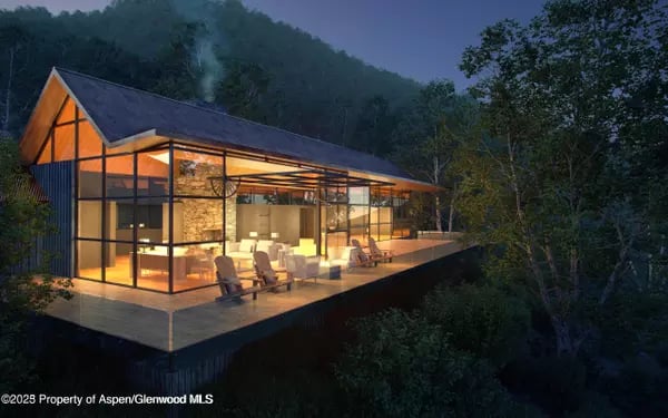 Modern glass-fronted home glowing at night, with a broad deck surrounded by wooded hills.