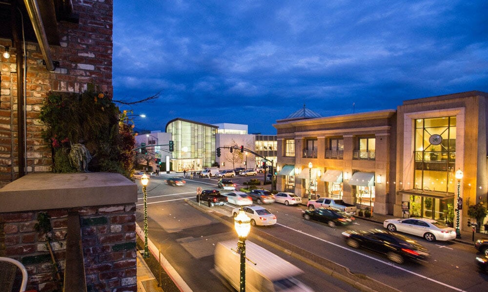 Downtown street at dusk with illuminated storefronts, brick buildings, and passing cars.