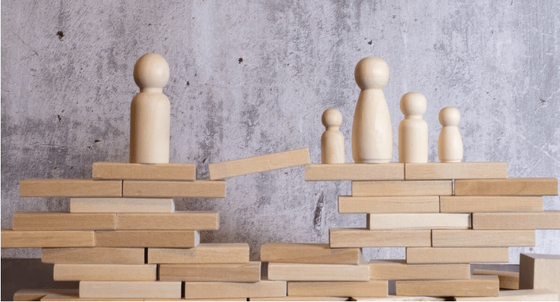 Wooden peg figures stand on block stacks linked by a narrow plank.