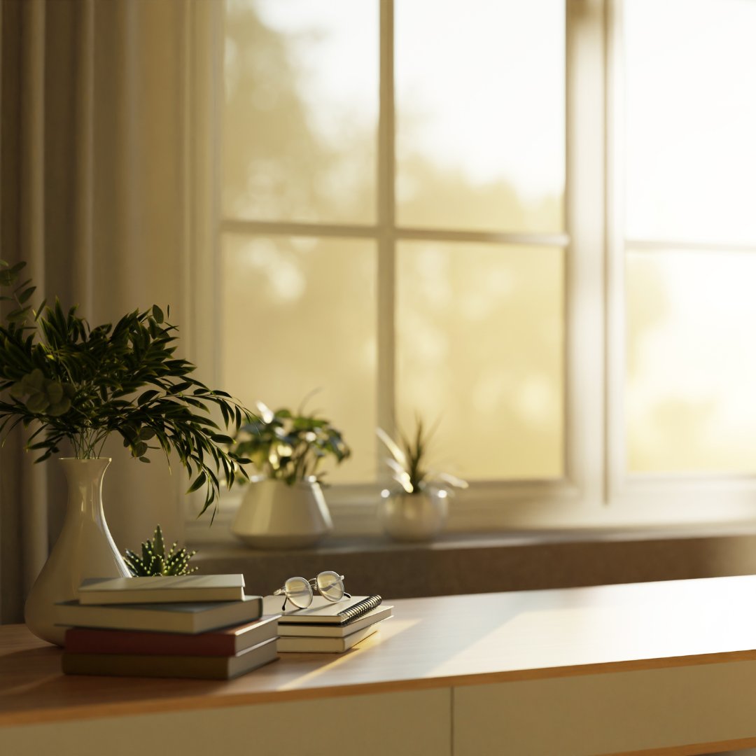 Sunlit desk by a large window with stacked books, eyeglasses, and potted plants.