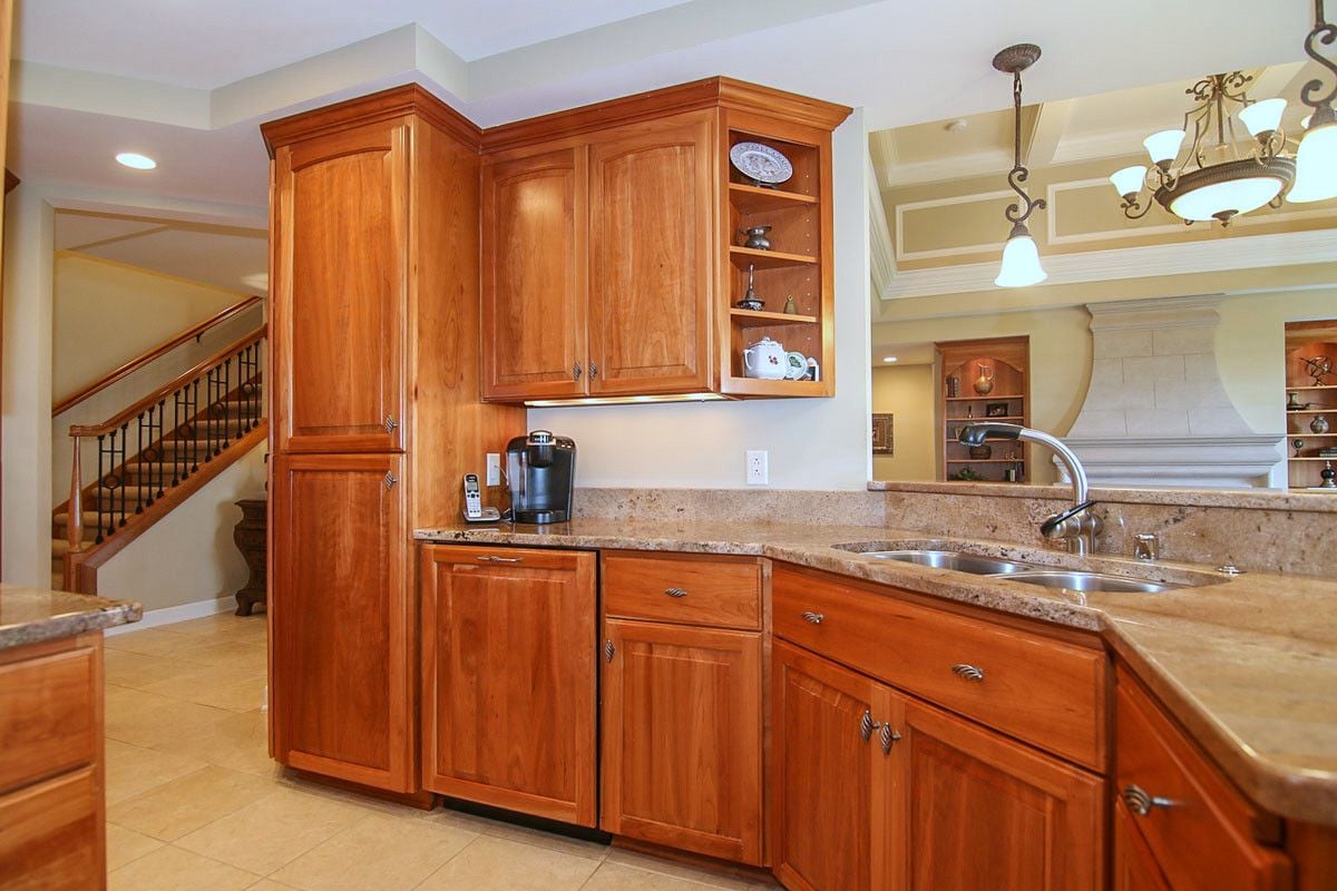 Kitchen with reddish wood cabinets, stone counters, and a double sink beside an open dining area.