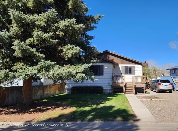 Single-story house with a raised wooden porch, shaded lawn, and large evergreen tree.