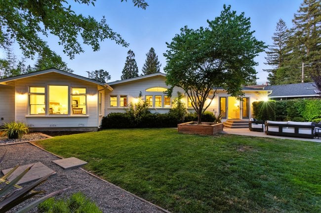 Single-story home at dusk, with a green lawn, mature trees, and patio seating.