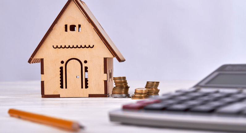 Wooden model house beside stacks of coins, a calculator, and a pencil.