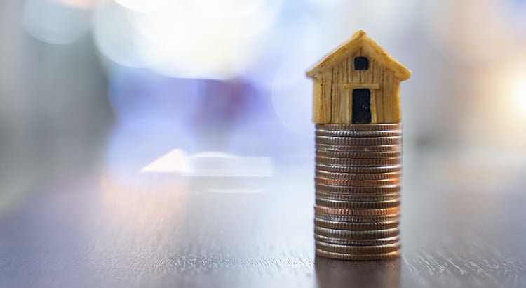 Small wooden house balanced atop a tall stack of coins against a blurred background.