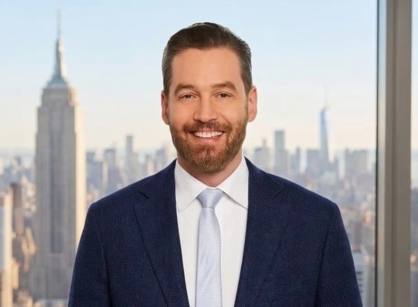 Smiling man in a navy suit and white tie against the New York skyline.