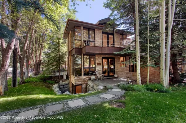 Two-story stone and wood house with a balcony and a stone path through a leafy yard.