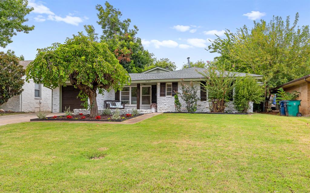 Single-story stone-front home with mature trees, a flower bed, and a broad lawn.