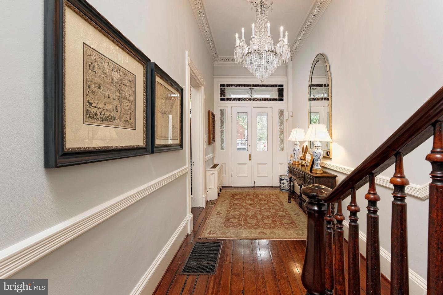 Elegant entry hall with a crystal chandelier, framed maps, hardwood floors, and a dark wood staircase.