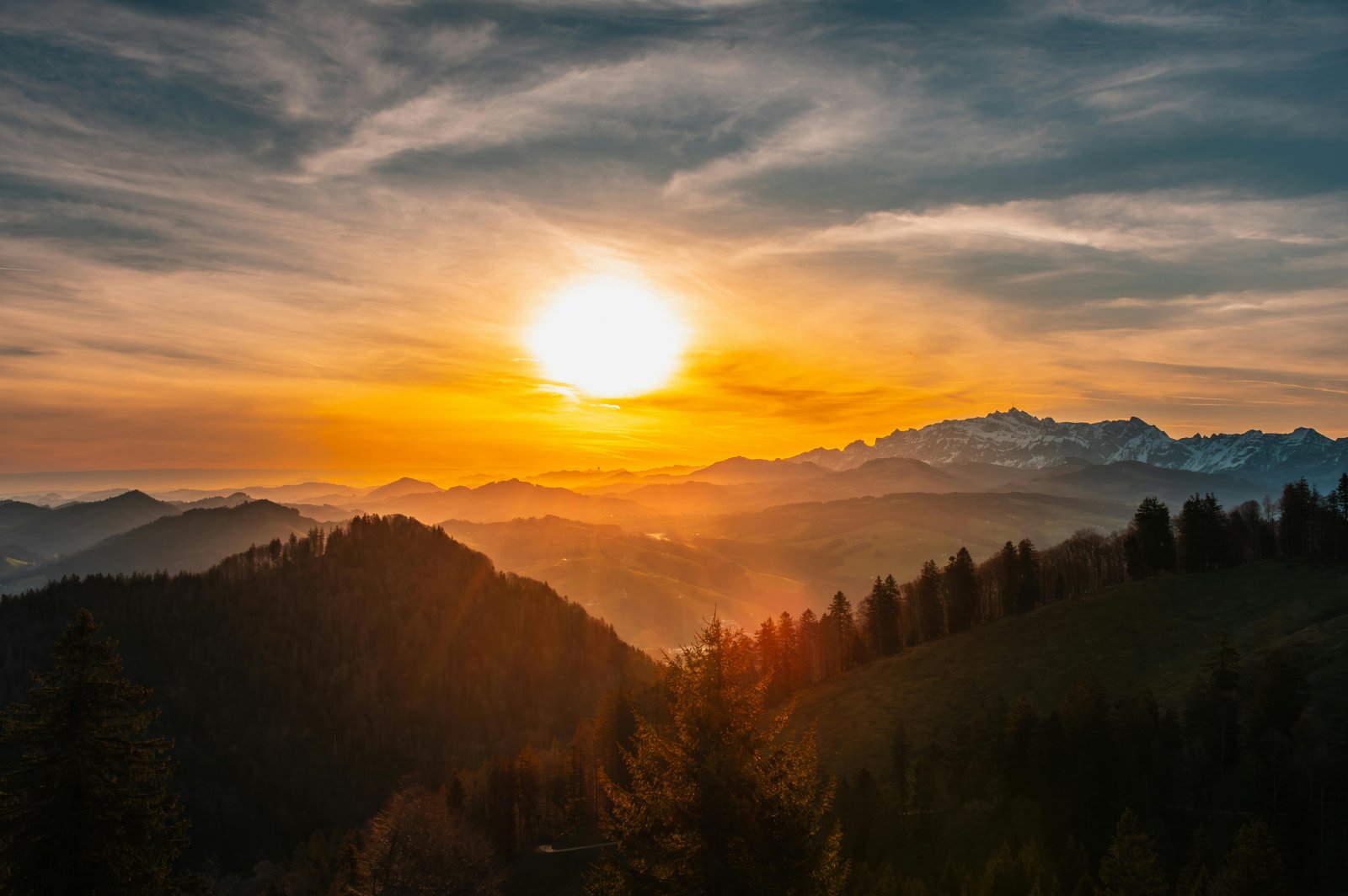 Sunset over forested mountain ridges, with a distant snow-dusted range beneath dramatic clouds.