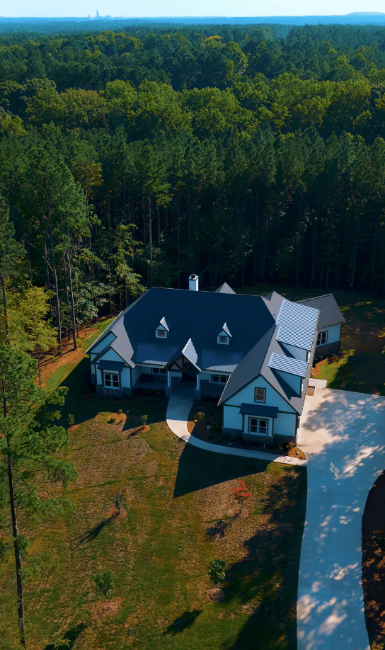 Aerial view of a large house with dark roofs and a curved driveway, surrounded by dense woods.