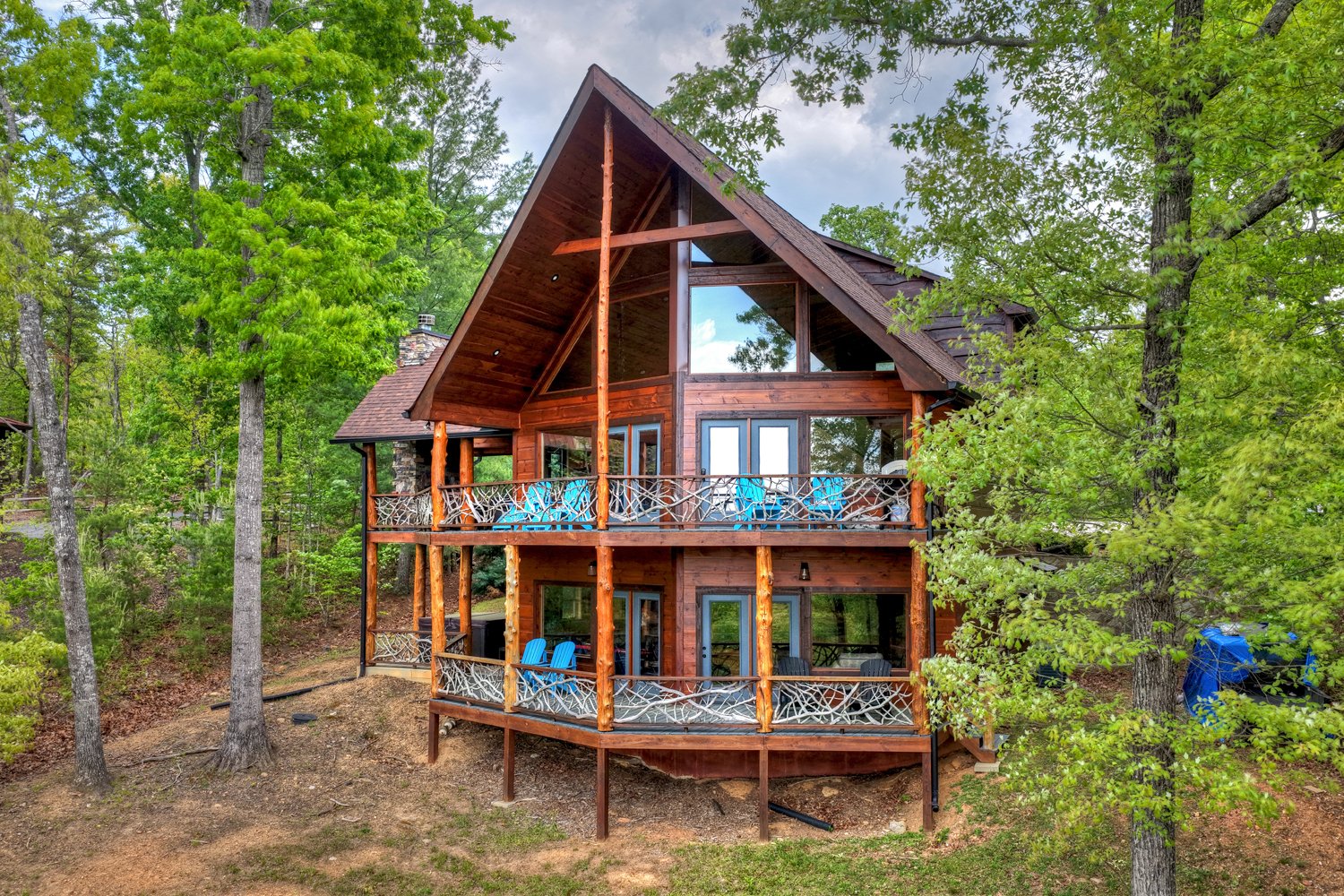 Timber A-frame mountain home with two balconies and blue Adirondack chairs among leafy trees.