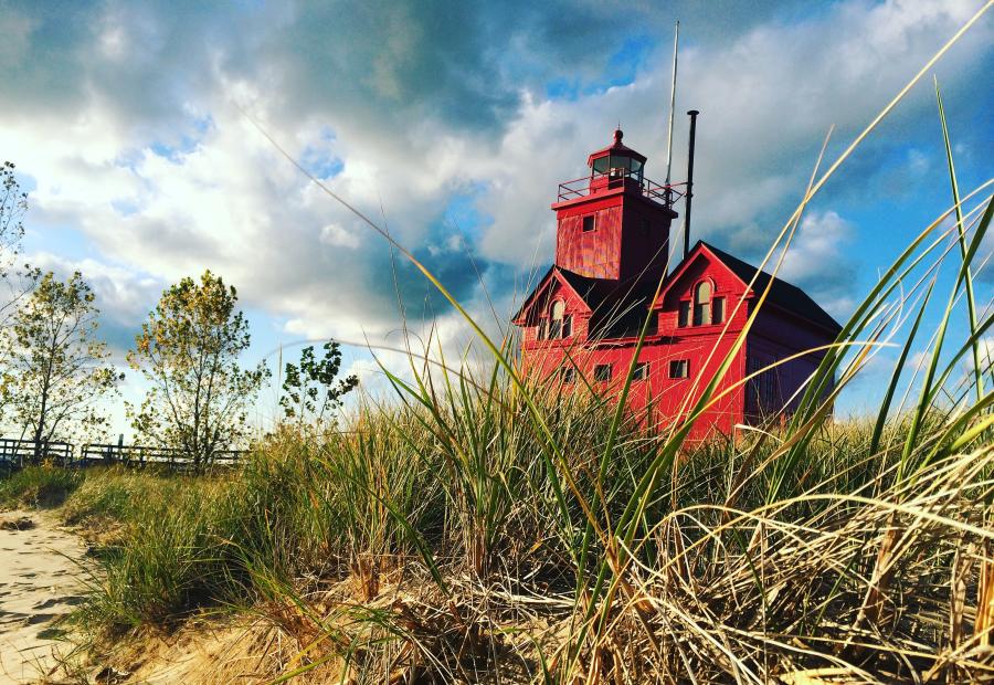 olland’s Big Red Lighthouse on Lake Michigan, viewed from the sandy beach with dune grass in the foreground.