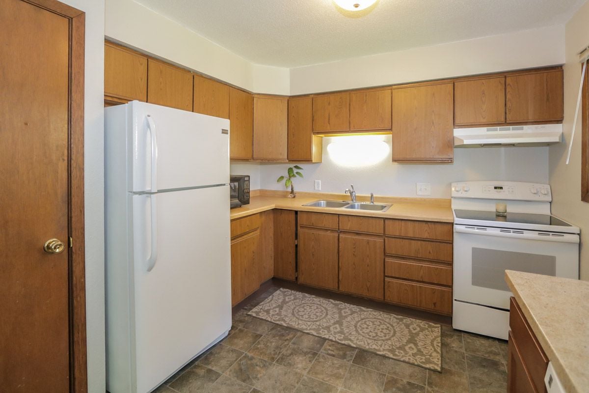 Kitchen with honey-colored cabinets, white appliances, a double-basin sink, and patterned floor mat.