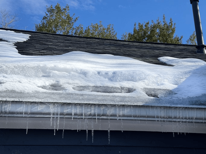 Snow-covered house roof with long icicles hanging from the gutter beneath a clear blue sky.