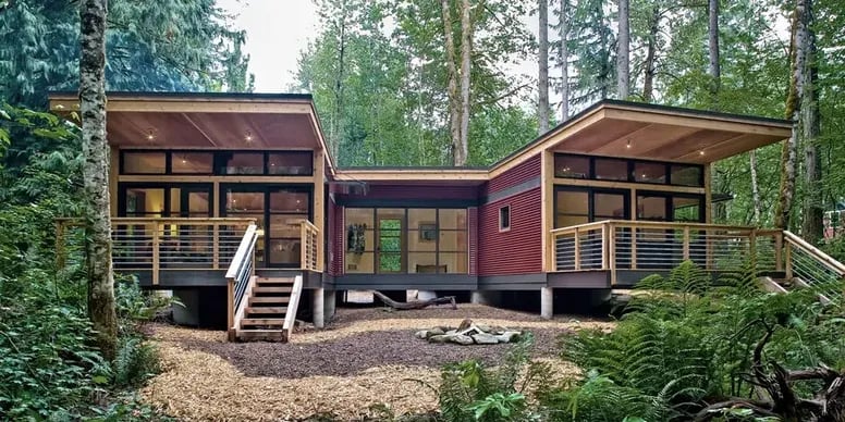 Modern modular home with red siding, raised decks, and broad roof overhangs among tall trees.