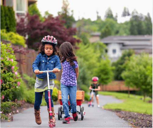Children ride scooters along a paved neighborhood path, with gardens and houses in the background.