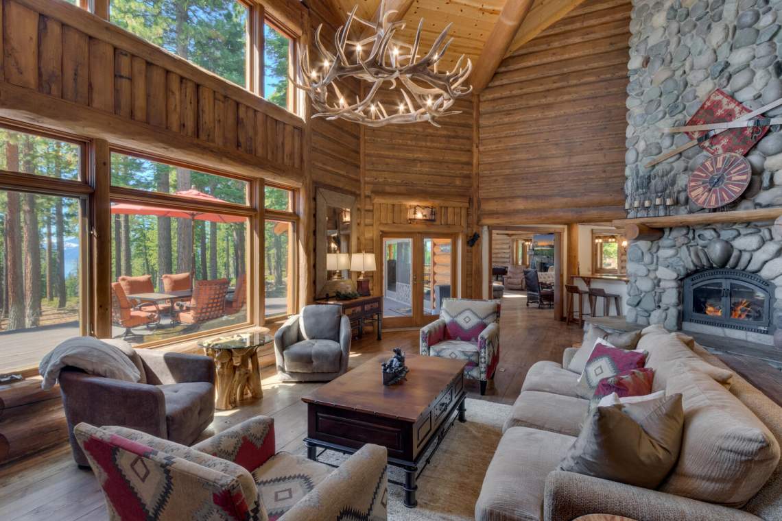 Timber living room with stone fireplace, antler chandelier, and windows overlooking trees and water.