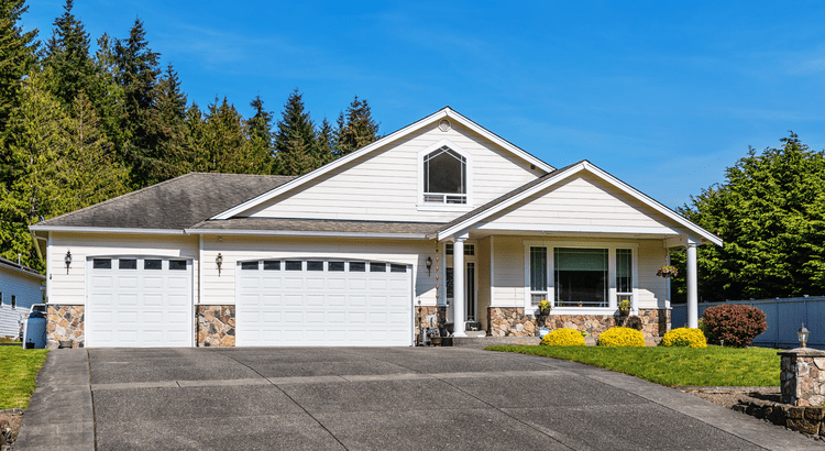 A white suburban house with a double garage and well-maintained front yard. Surrounded by tall trees under a clear blue sky, conveying a peaceful vibe.