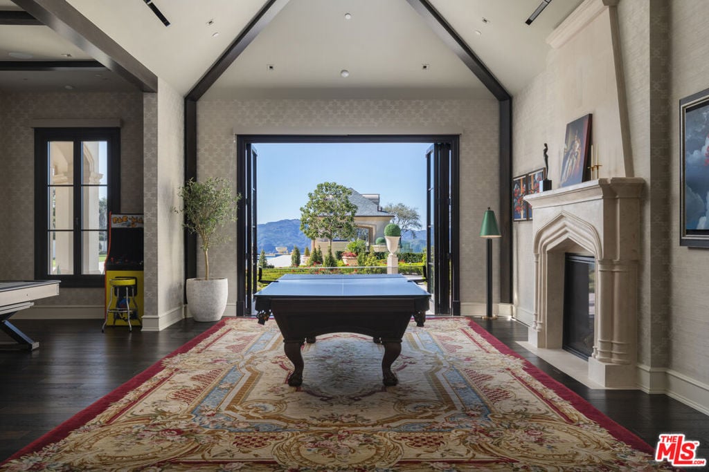 Billiards room with a pool table, ornate rug, stone fireplace, and open doors framing landscaped grounds and mountains.