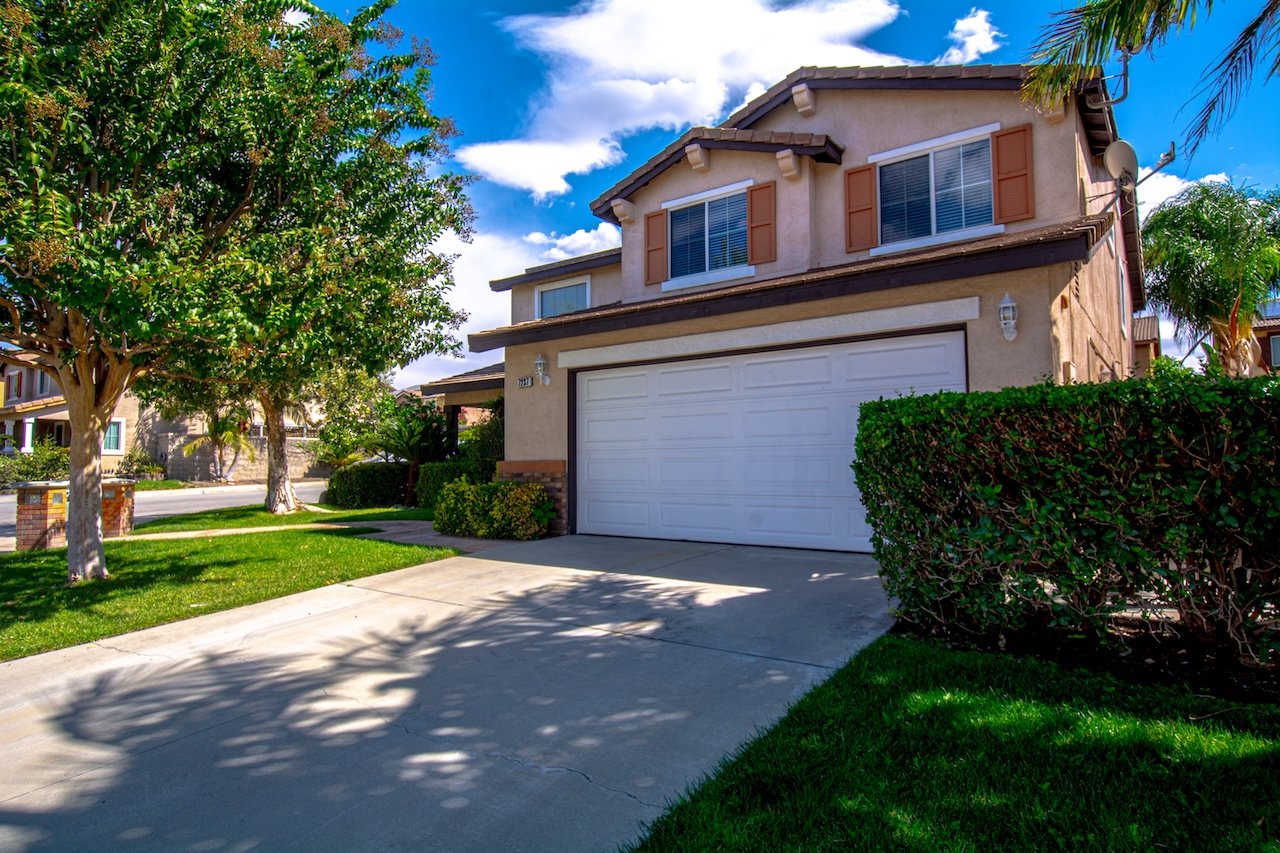 Two-story beige stucco house with a white double garage, broad driveway, hedges, and mature trees.