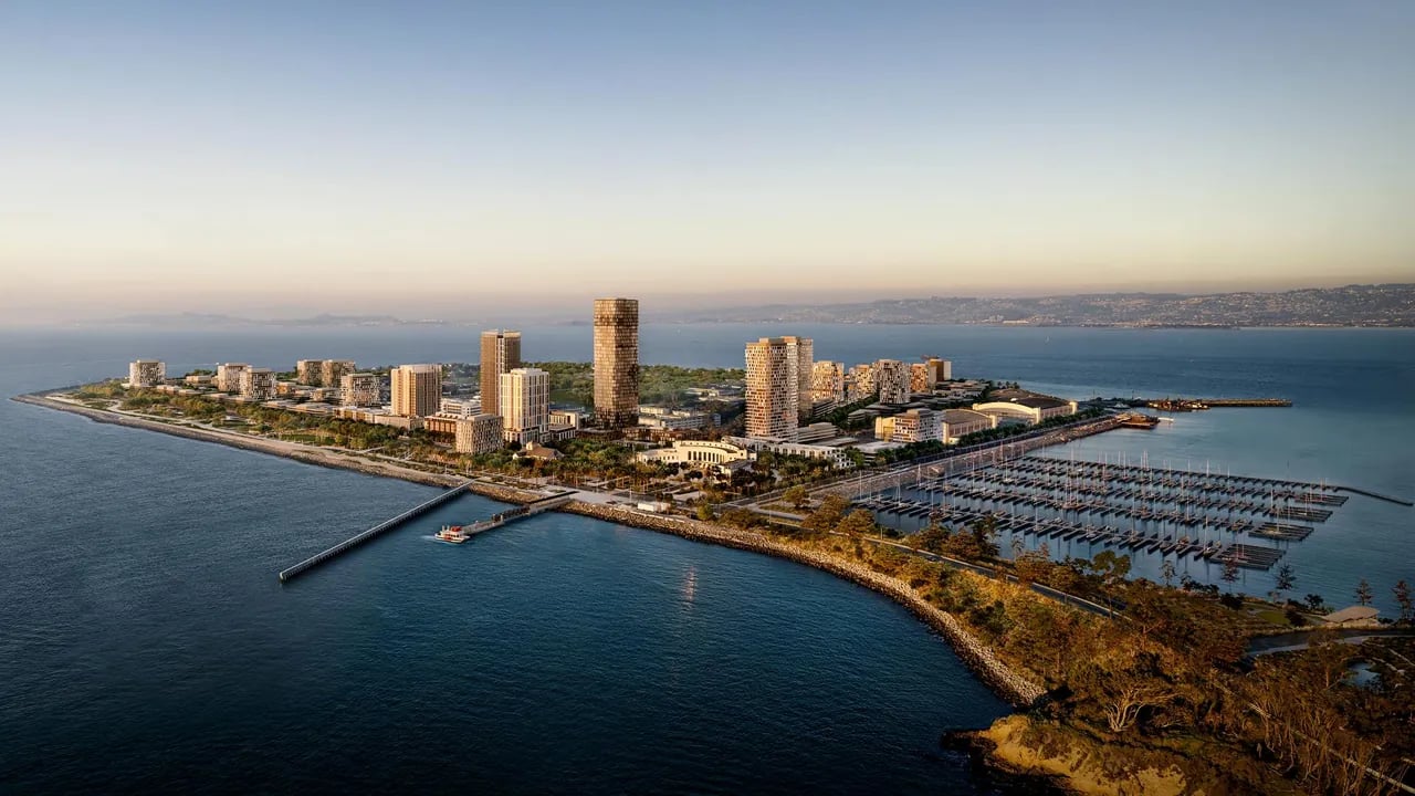 Aerial view of Treasure Island showing modern high-rise residential towers, marina with boats, pier, and San Francisco Bay with distant hills under golden hour sky