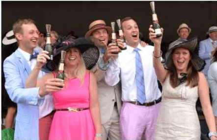 Festival attendees celebrate with raised wine bottles; two women wear pink and white outfits.