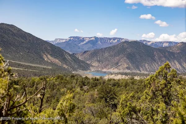 Mountain valley with scrub-covered hillsides, distant water, and a partly cloudy blue sky.