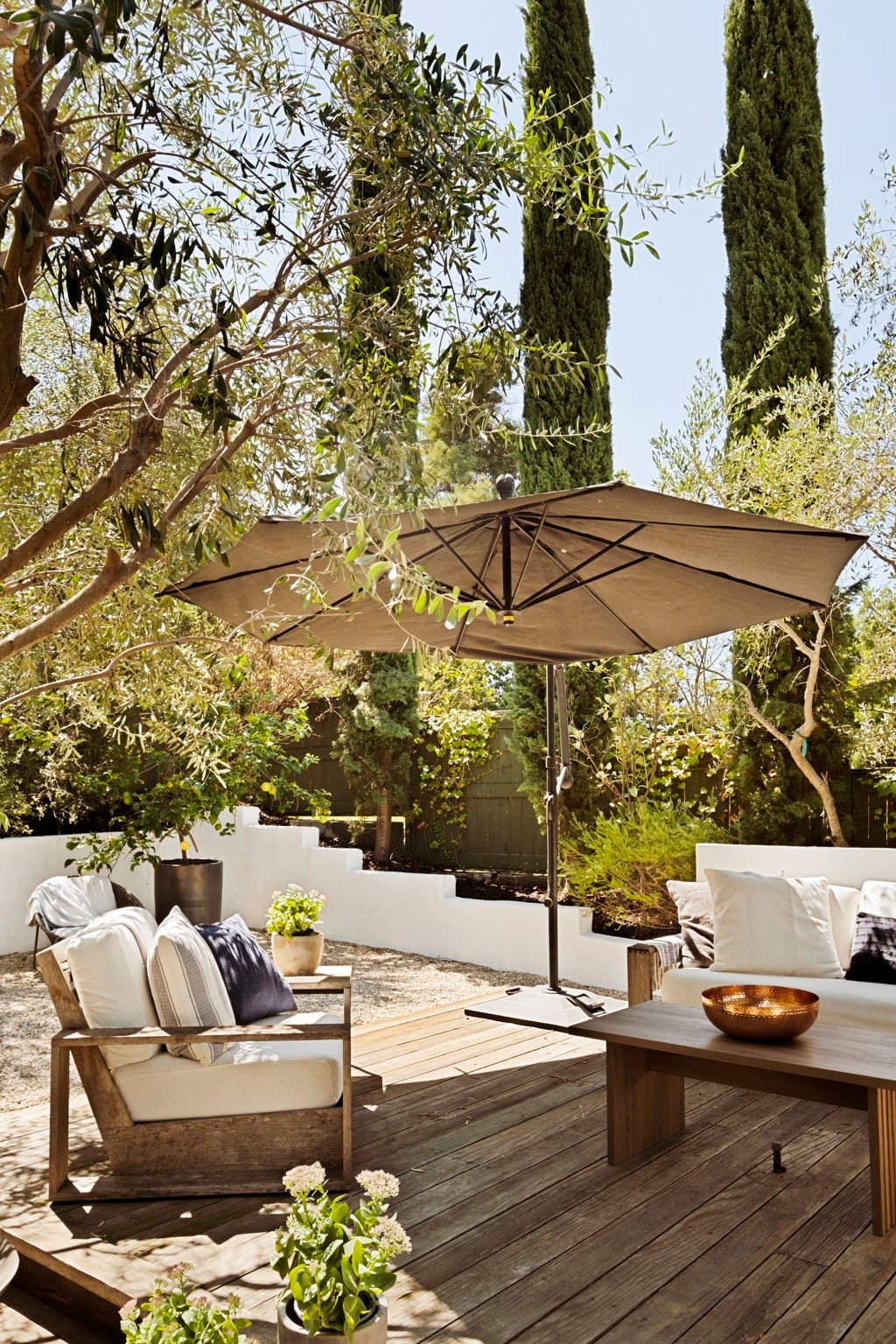 Outdoor patio with wooden deck, cushioned seating, large umbrella, and tall cypress trees.