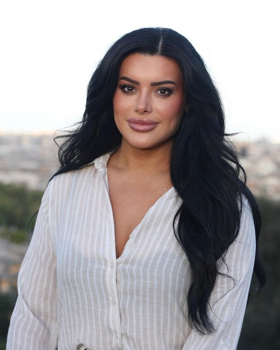 Biana Xherimeja in a white striped blouse, with long dark hair and a softly blurred city background.