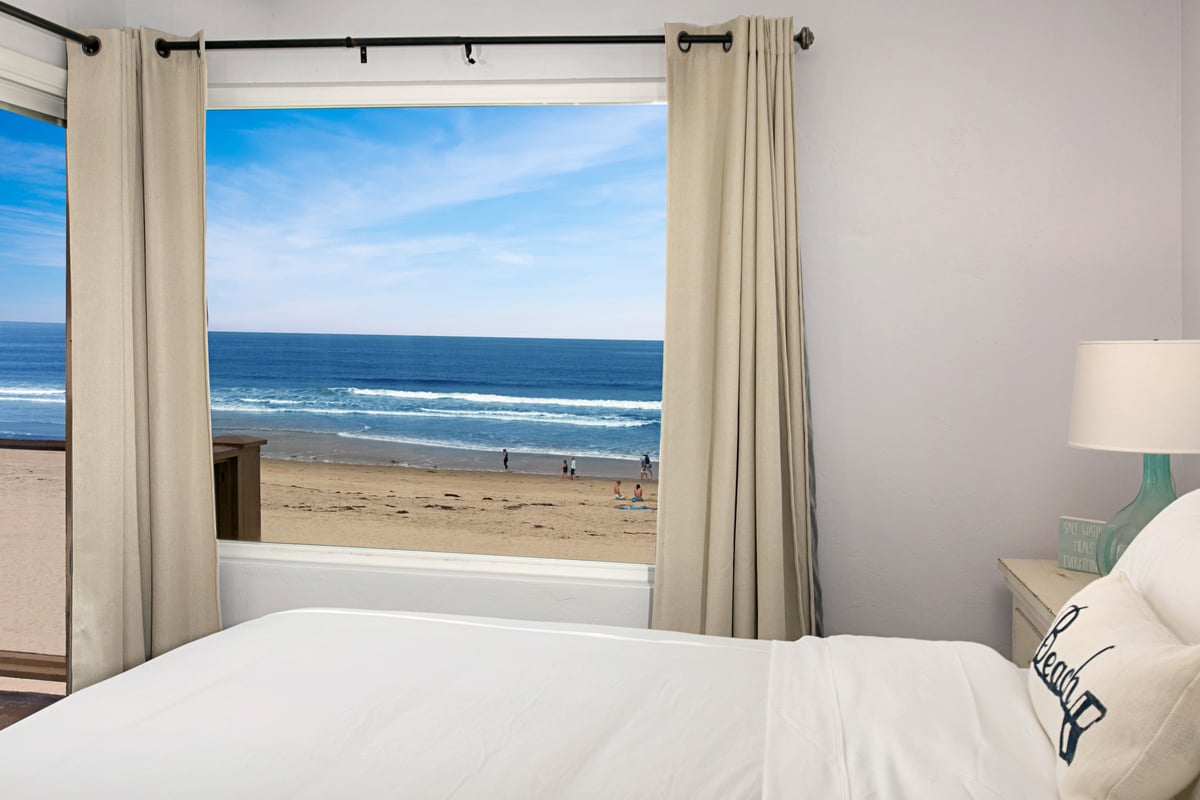 Bedroom with cream curtains framing a view of ocean waves and a sandy beach.