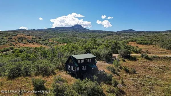 Dark two-story house amid open brushland, trees, and distant mountain ridges.