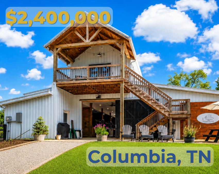 Barn-style property with a wood balcony and staircase, outdoor seating, and a green lawn.
