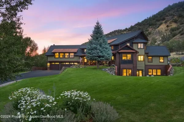 Multi-level home with glowing windows, a tall evergreen, and a green hillside lawn at dusk.