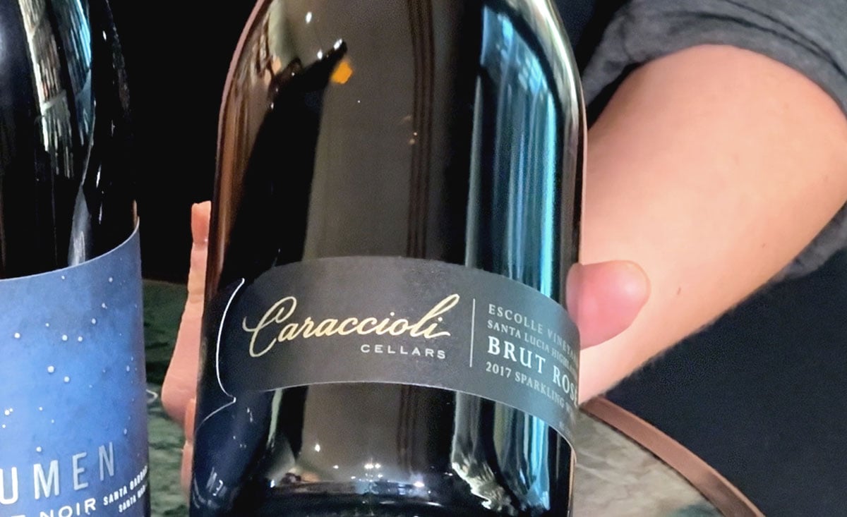 A hand holding a bottle of Caraccioli Cellars Brut Rosé sparkling wine, with another wine bottle partially visible in the background.