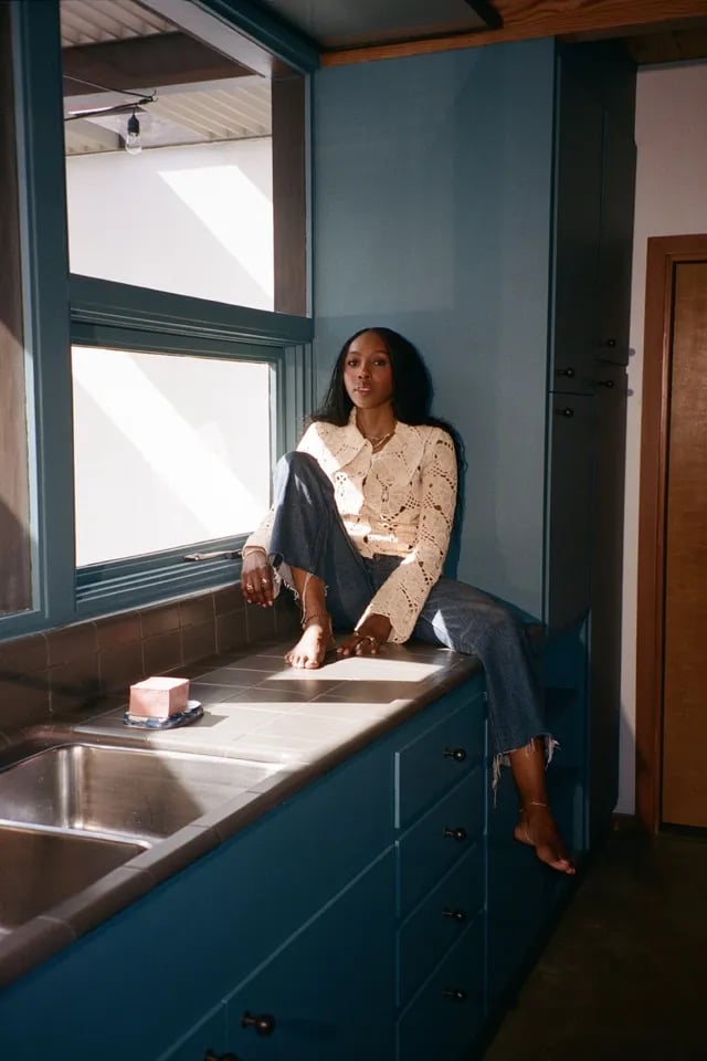 Aiesha Bailey seated barefoot on a blue kitchen counter beside a large sunlit window.
