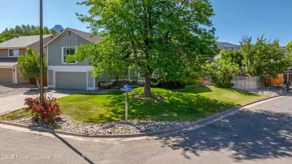 Gray two-story home with an attached garage, shaded front lawn, and large mature tree.