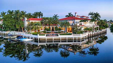 Waterfront building with red tile roofs, palm trees, docks, and boats beside a calm canal.