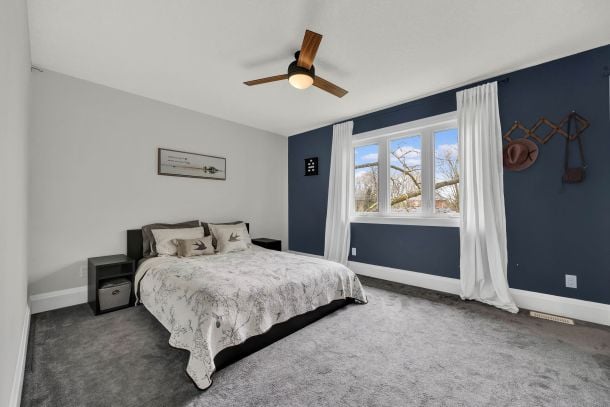 Bedroom with a navy accent wall, large curtained window, gray carpet, and wood-blade ceiling fan.