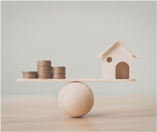 A miniature house and coin stacks balanced on a plank above a wooden sphere
